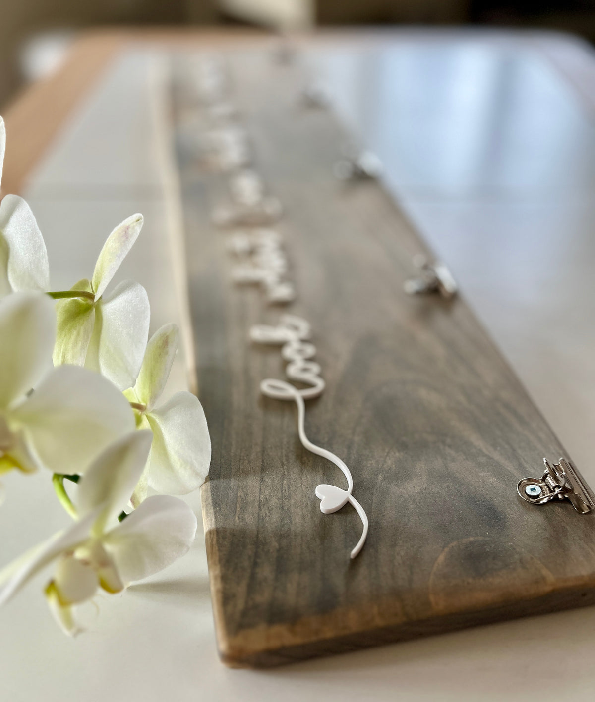 Wooden art display sign with white cursive lettering and heart detail, natural wood grain background with clip holders on white table