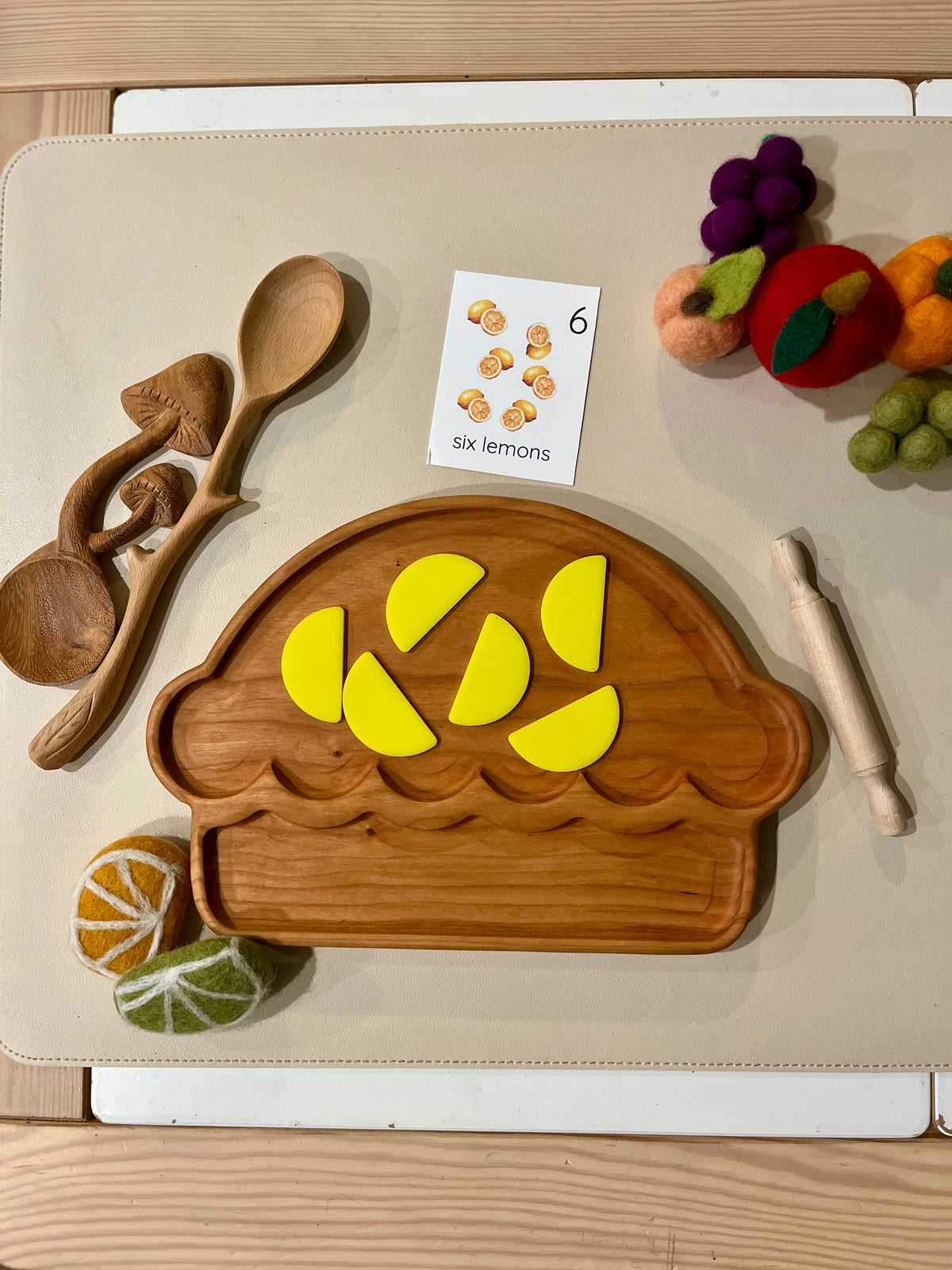 Wooden muffin-shaped tray with six bright yellow lemon wedge pieces, wooden utensils, felt fruit, and a counting card nearby