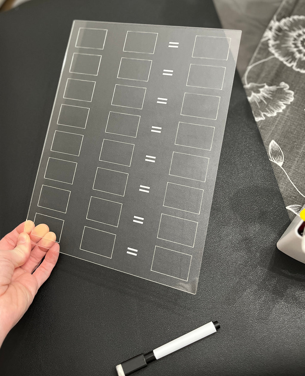Hand holding a clear acrylic board with equal sign and rectangular grids above a black surface with a black dry erase marker nearby