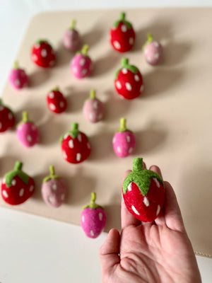 Hand holding a red felt strawberry with green leaves among scattered red and pink felt strawberries on a beige mat