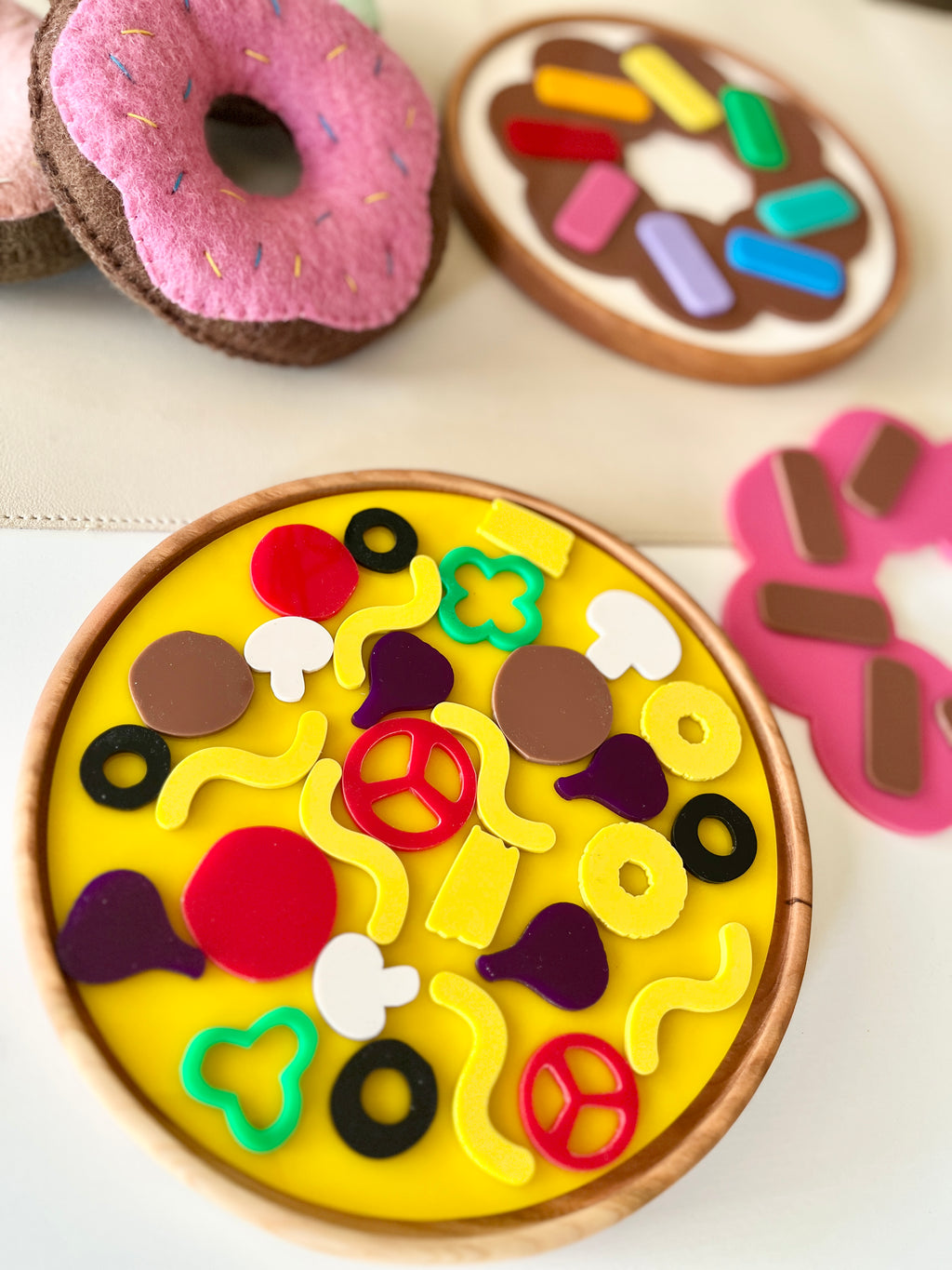 Wooden toy pizza with colorful toppings next to pink felt donut toys on a light surface