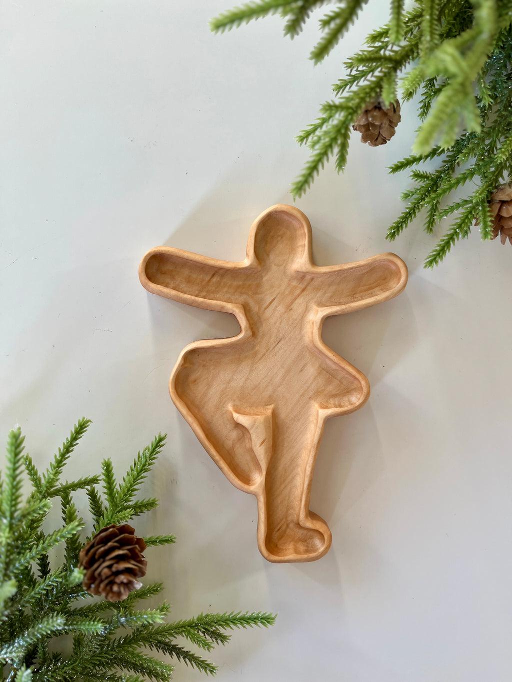 Carved wooden tray shaped like a dancing figure surrounded by pine branches and pinecones on white surface