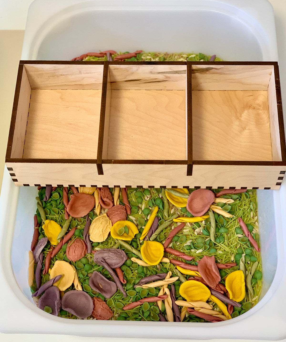 Wooden three-compartment tray positioned above a sensory bin filled with colorful dried pasta and green split peas