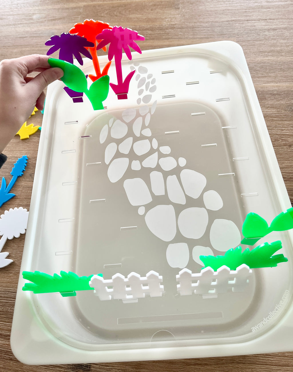 White tray with a printed stone pathway and slots, holding colorful translucent acrylic pegs shaped like plants and flowers being arranged by hand, featuring STEM Insert – Acrylic Insert with Different Peg Options - Neighborhood, Veggie Garden, Flower Garden, Under the Sea + More, handcrafted, homeschool, education resources.