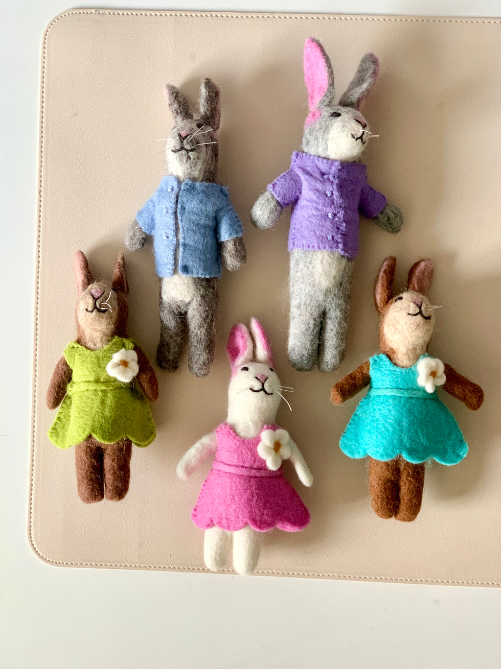 Five handmade felt bunny dolls wearing colorful clothes arranged on a beige surface