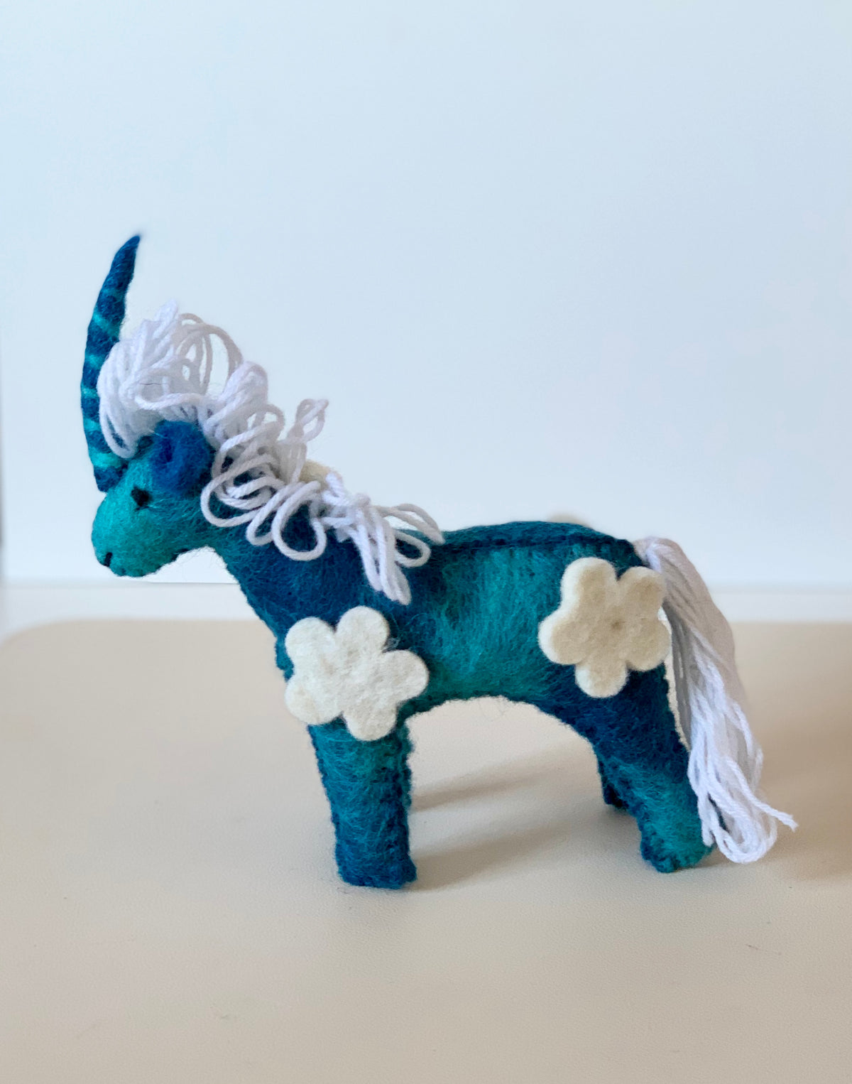 Small teal and navy felt unicorn with curly white yarn mane and tail, adorned with cream felt flower patches on beige surface
