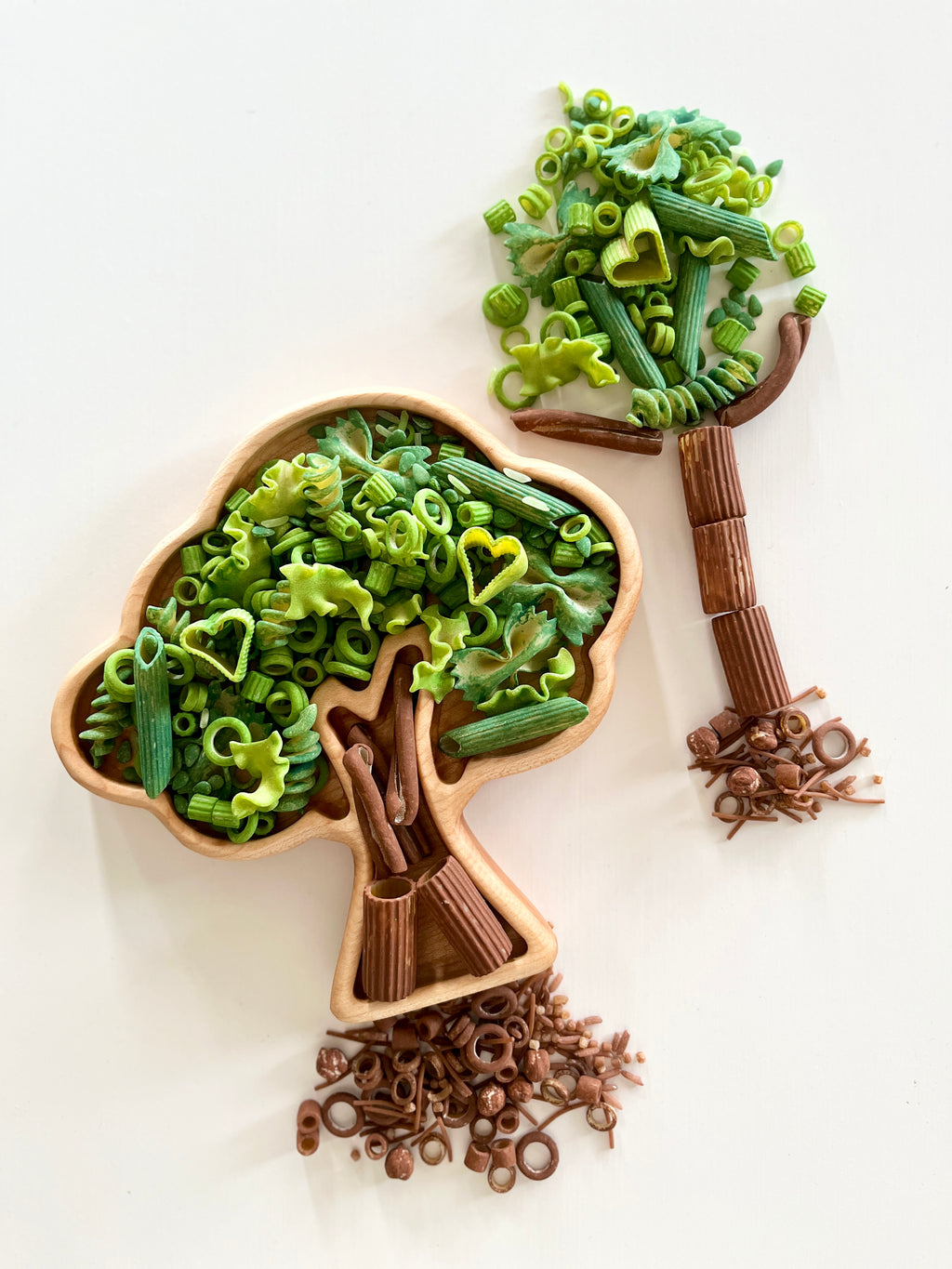 Assortment of green and brown pasta shapes arranged inside and outside a tree-shaped wooden tray on a white background