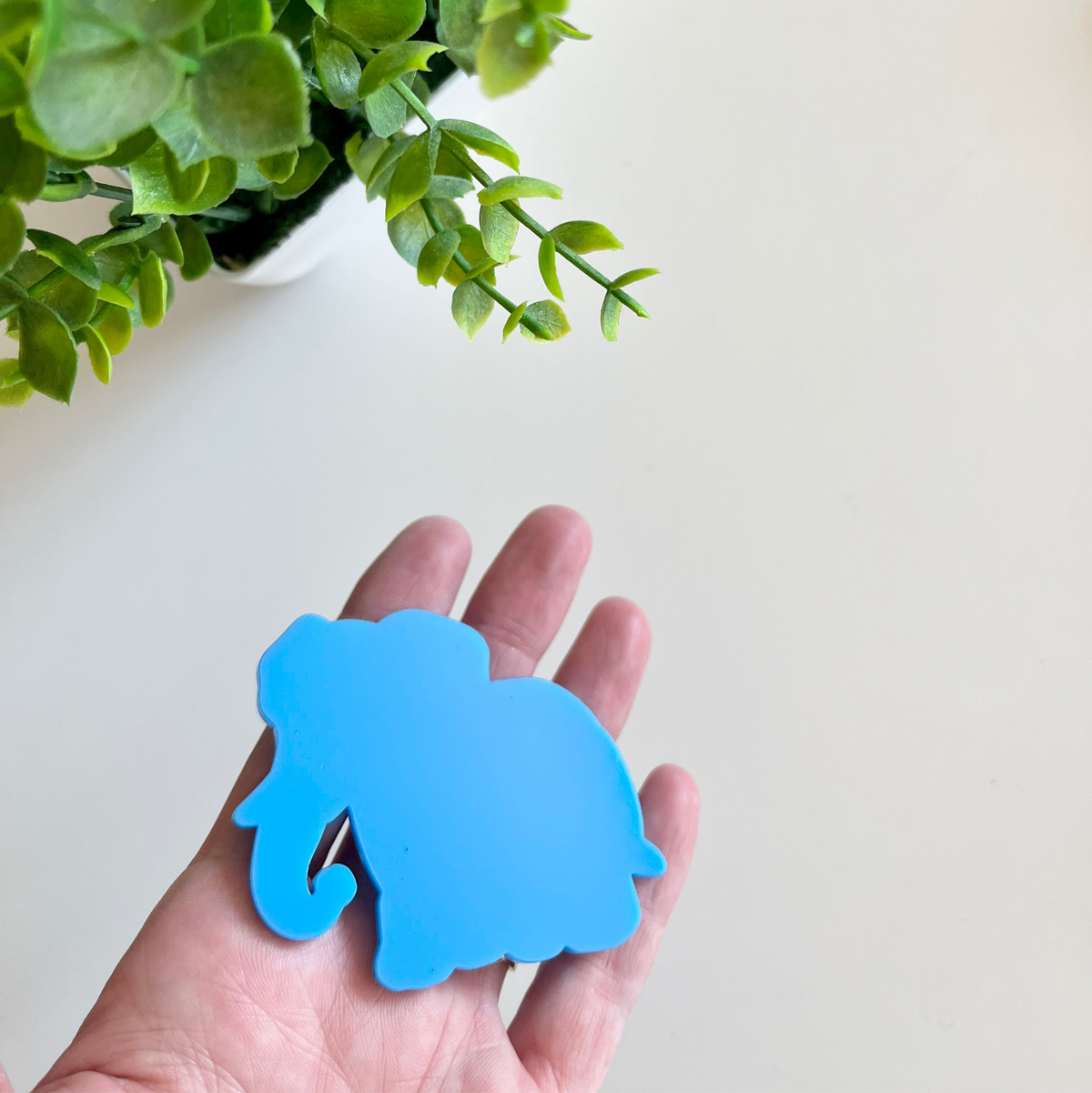 Hand holding a smooth blue elephant-shaped cutout with green leaves of a plant in the top corner on a light background