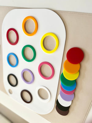 White board with colorful plastic rings arranged in rows and matching solid discs stacked beside it on a beige surface