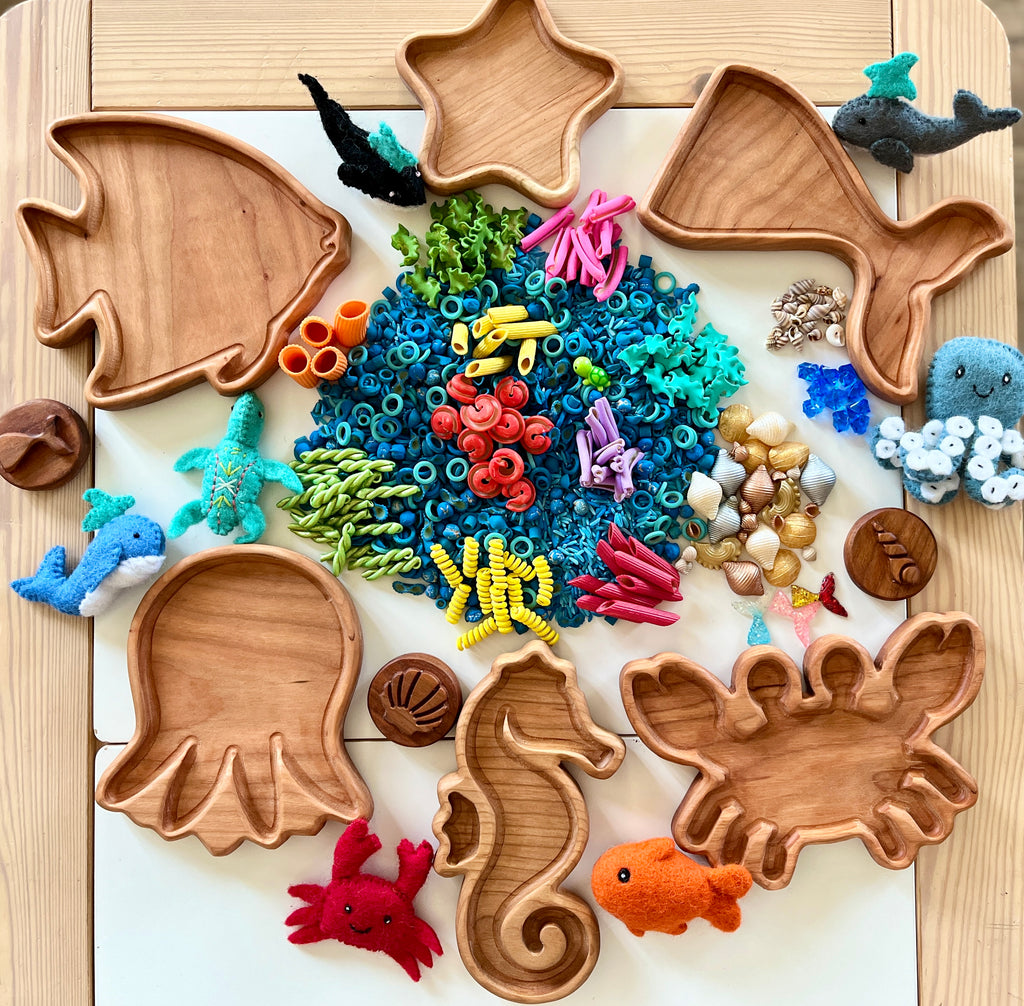 Wooden sea creature-shaped trays with colorful sensory pasta, shells, and felt marine animals arranged on a light surface.
