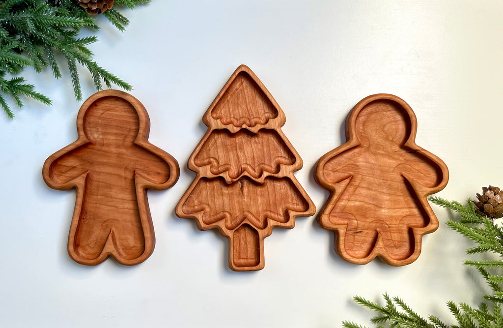 Three carved wooden trays shaped like gingerbread man, Christmas tree, and gingerbread woman with pine sprigs nearby
