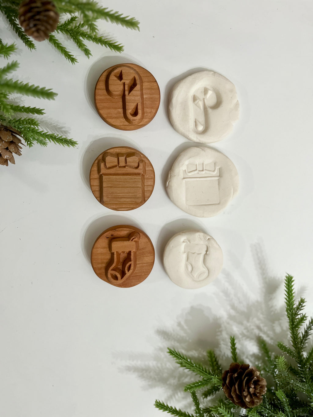 Three wooden holiday stamps with candy cane, gift box, and Christmas stocking alongside white clay imprints on white surface with pine branches and cones