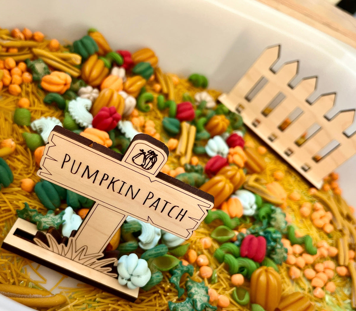Miniature colorful pumpkin-shaped decorations and small gourds scattered on yellow shredded material with a wooden "Pumpkin Patch" sign and fence