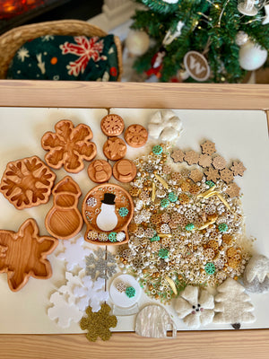Wooden snowman and snowflake-shaped trays alongside natural and craft sensory materials on white tabletop near decorated Christmas tree