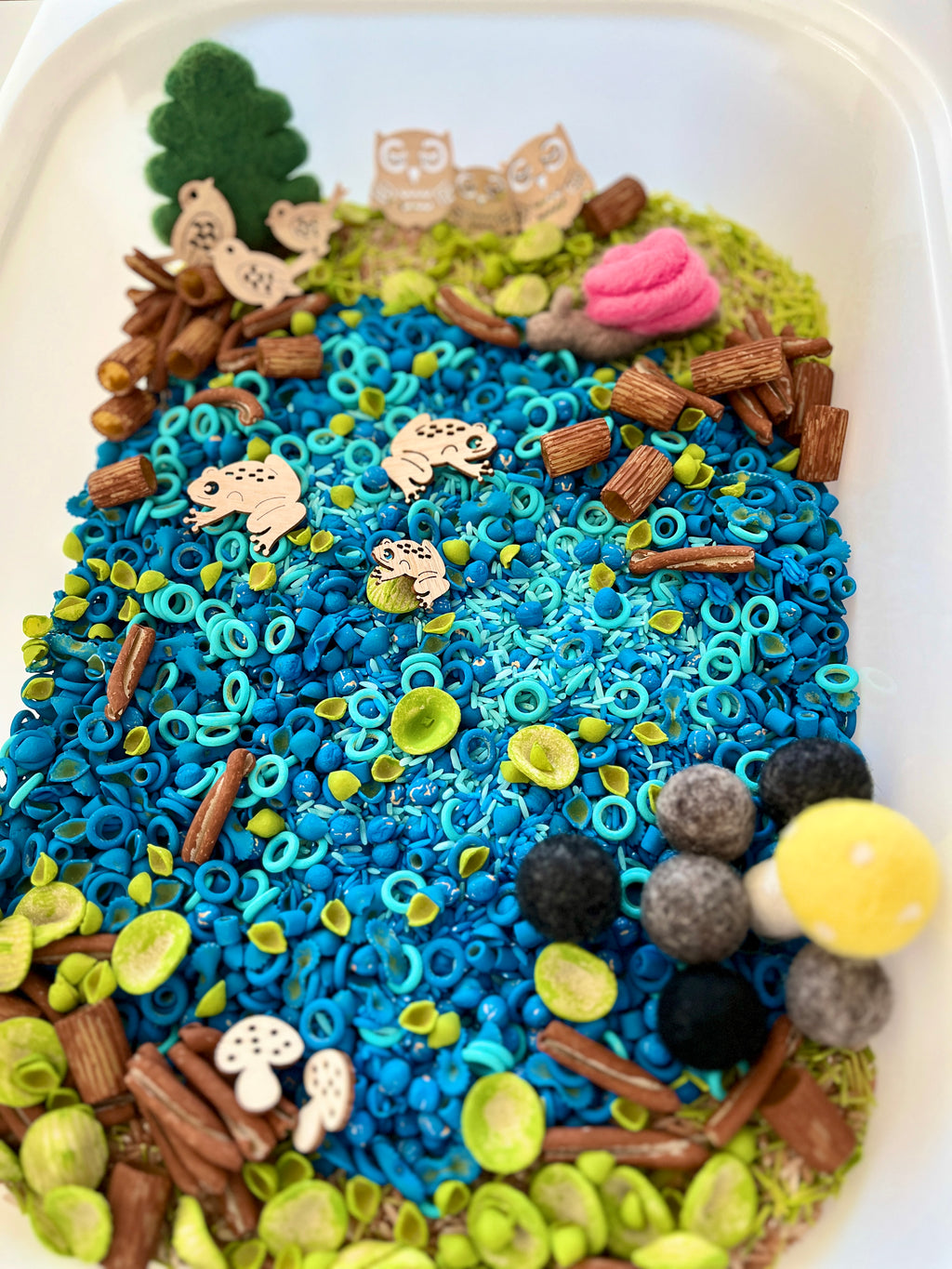 Sensory tray with blue pasta and felt balls resembling a pond, wooden animal shapes, green moss, and small sticks scattered around.