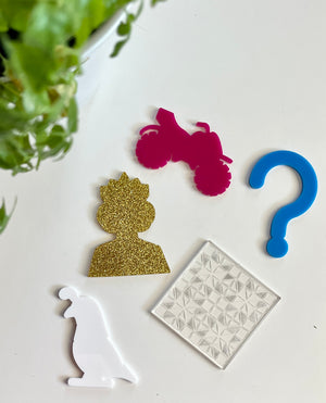 Five acrylic shapes including gold glitter silhouette, pink motorcycle, blue question mark, white bird, and clear textured square on white surface