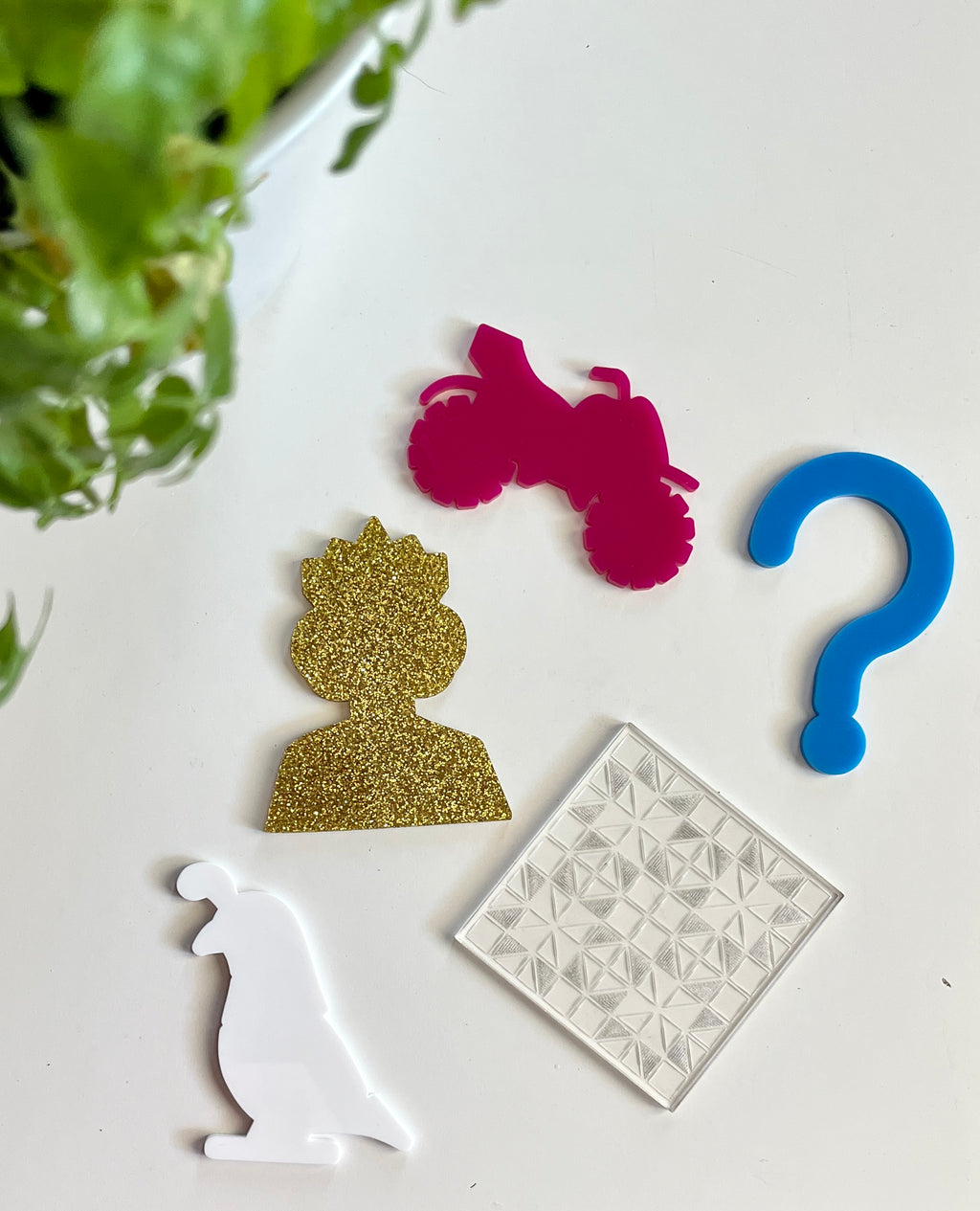 Five acrylic shapes including gold glitter silhouette, pink motorcycle, blue question mark, white bird, and clear textured square on white surface