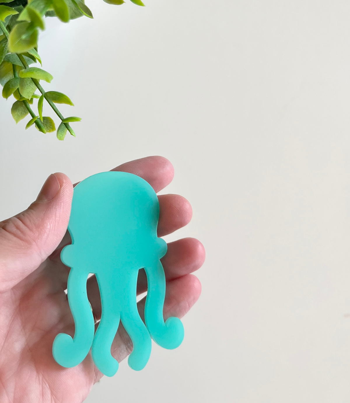 Hand holding a smooth turquoise octopus-shaped cutout with green leafy plant leaves above on a light background