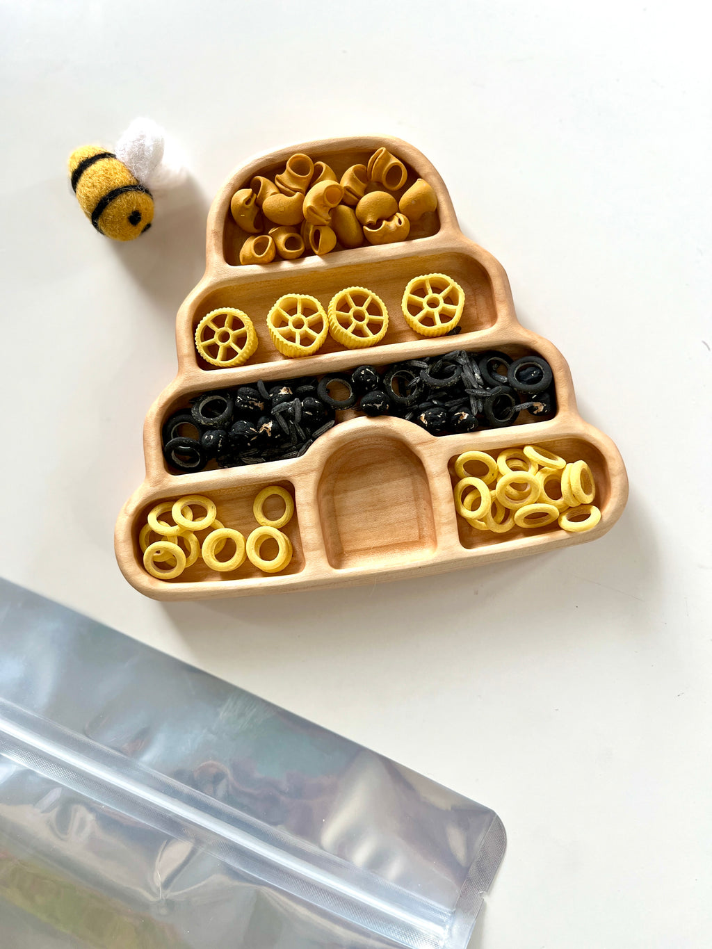Wooden beehive tray with yellow and black pasta shapes beside a small felt bee on white surface