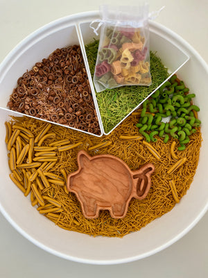White segmented tray filled with brown, yellow, and green pasta shapes, with a wooden pig scoop and small bag of multicolored pasta