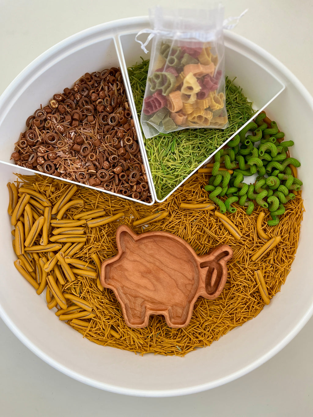 White segmented tray filled with brown, yellow, and green pasta shapes, with a wooden pig scoop and small bag of multicolored pasta