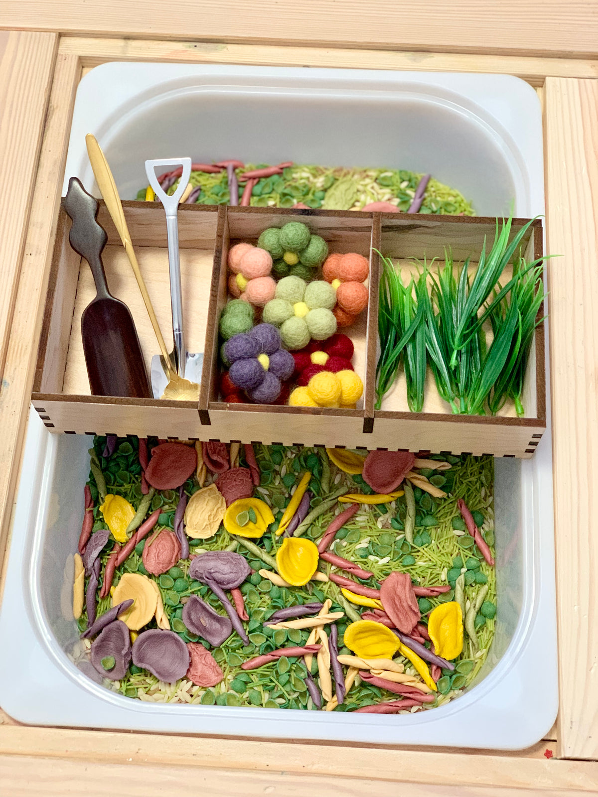 Toy gardening tools and felt flowers arranged in a wooden tray above colorful dried pasta and green seeds in a sensory bin