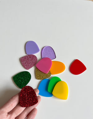 Hand holding a red glitter plastic gumdrop piece with a collection of colorful smooth and glittered plastic gumdrops in purple, pink, orange, gold, green, blue, and yellow on a white surface, Little Dry Erasables — Set of 12 Gumdrops, handcrafted homeschool educational resources.