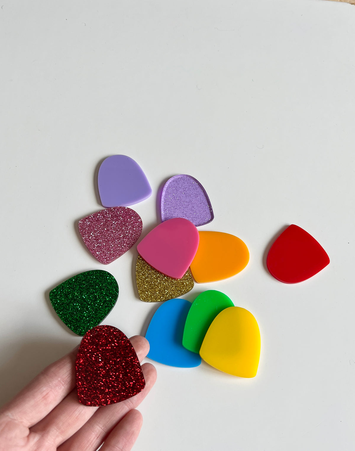 Hand holding a red glitter plastic gumdrop piece with a collection of colorful smooth and glittered plastic gumdrops in purple, pink, orange, gold, green, blue, and yellow on a white surface, Little Dry Erasables — Set of 12 Gumdrops, handcrafted homeschool educational resources.