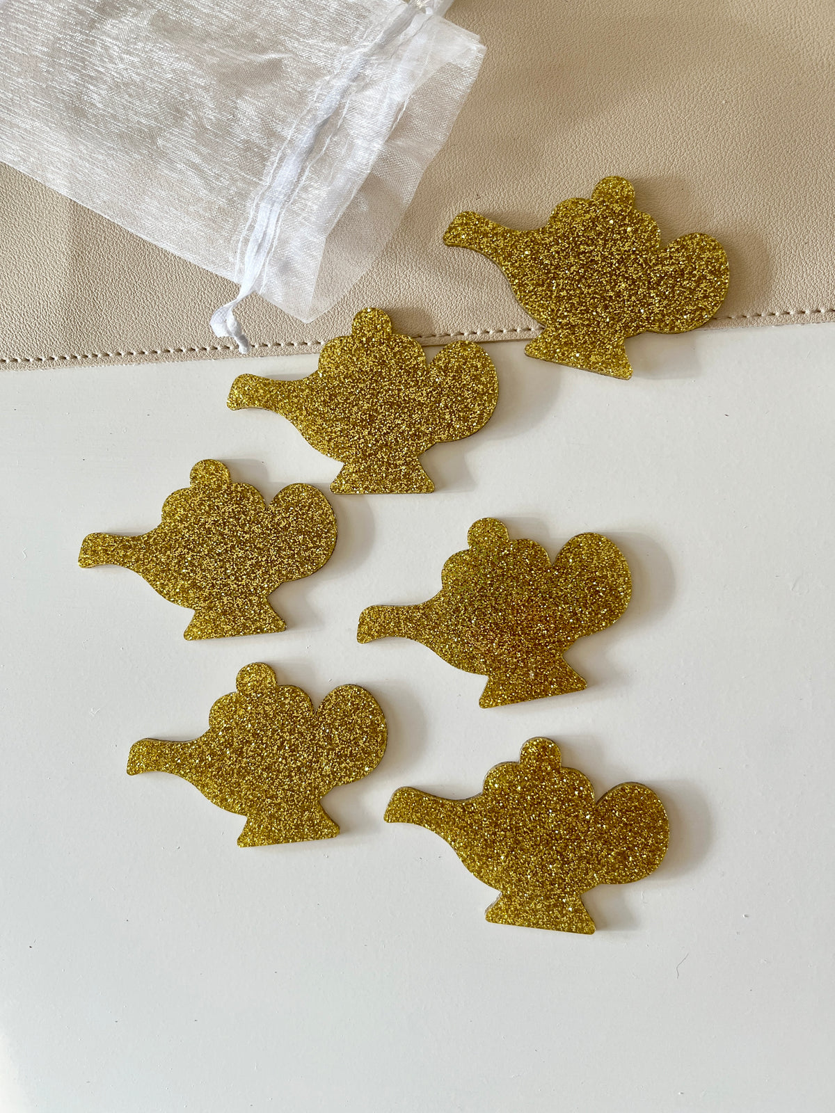 Six gold glitter genie lamp-shaped pieces arranged near a white sheer drawstring pouch on a two-tone surface