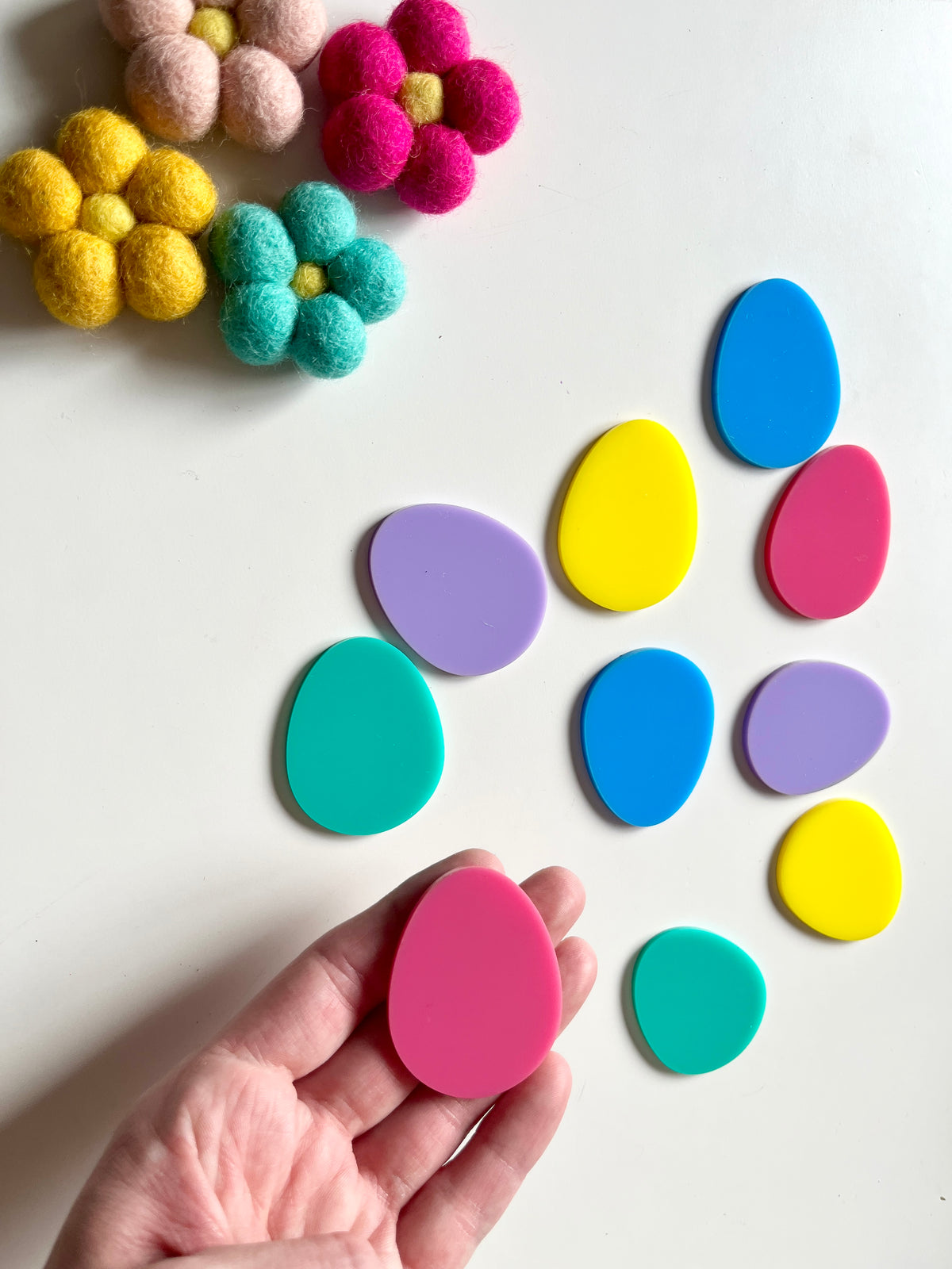 Hand holding pink egg-shaped plastic piece surrounded by scattered colorful pastel egg shapes and felt flowers on white surface