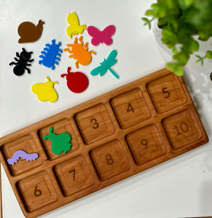 Wooden tray with numbered slots 1 to 10 and colorful insect and bug-shaped cutouts on white surface next to green plant