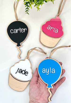 Four wooden name tags in balloon and cupcake shapes with black, pink, white, and blue colors and personalized white letters