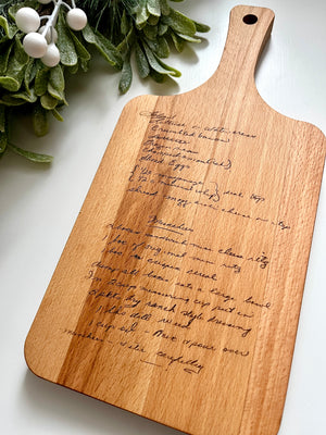 Natural wooden serving board with personalized handwritten recipe laser engraved and leafy green decor on white background
