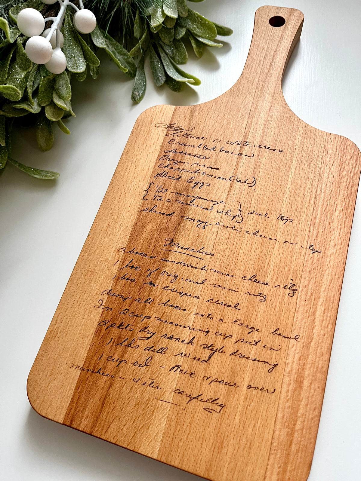 Natural wooden serving board with personalized handwritten recipe laser engraved and leafy green decor on white background