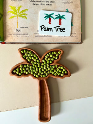 Wooden palm tree-shaped tray filled with green peas placed on a beige surface below an open book about palm trees