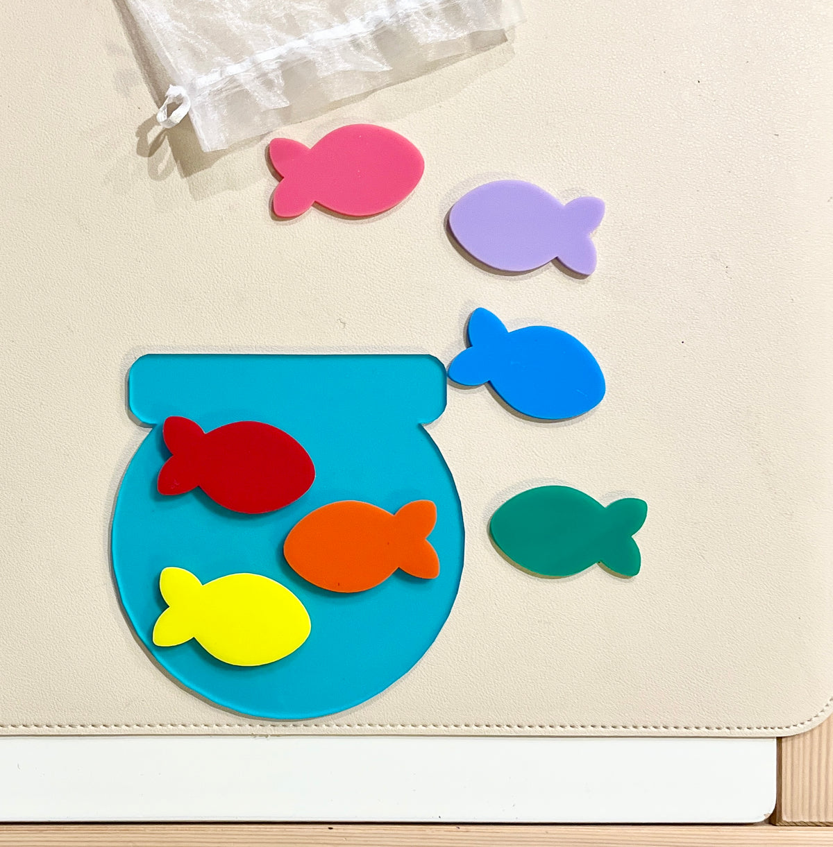 Colorful fish-shaped puzzle pieces with a blue fishbowl on a beige surface next to a white drawstring bag