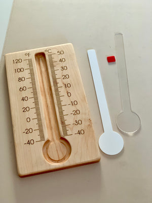 Wooden thermometer board with Fahrenheit and Celsius scales next to separate clear and white plastic thermometer inserts on light surface