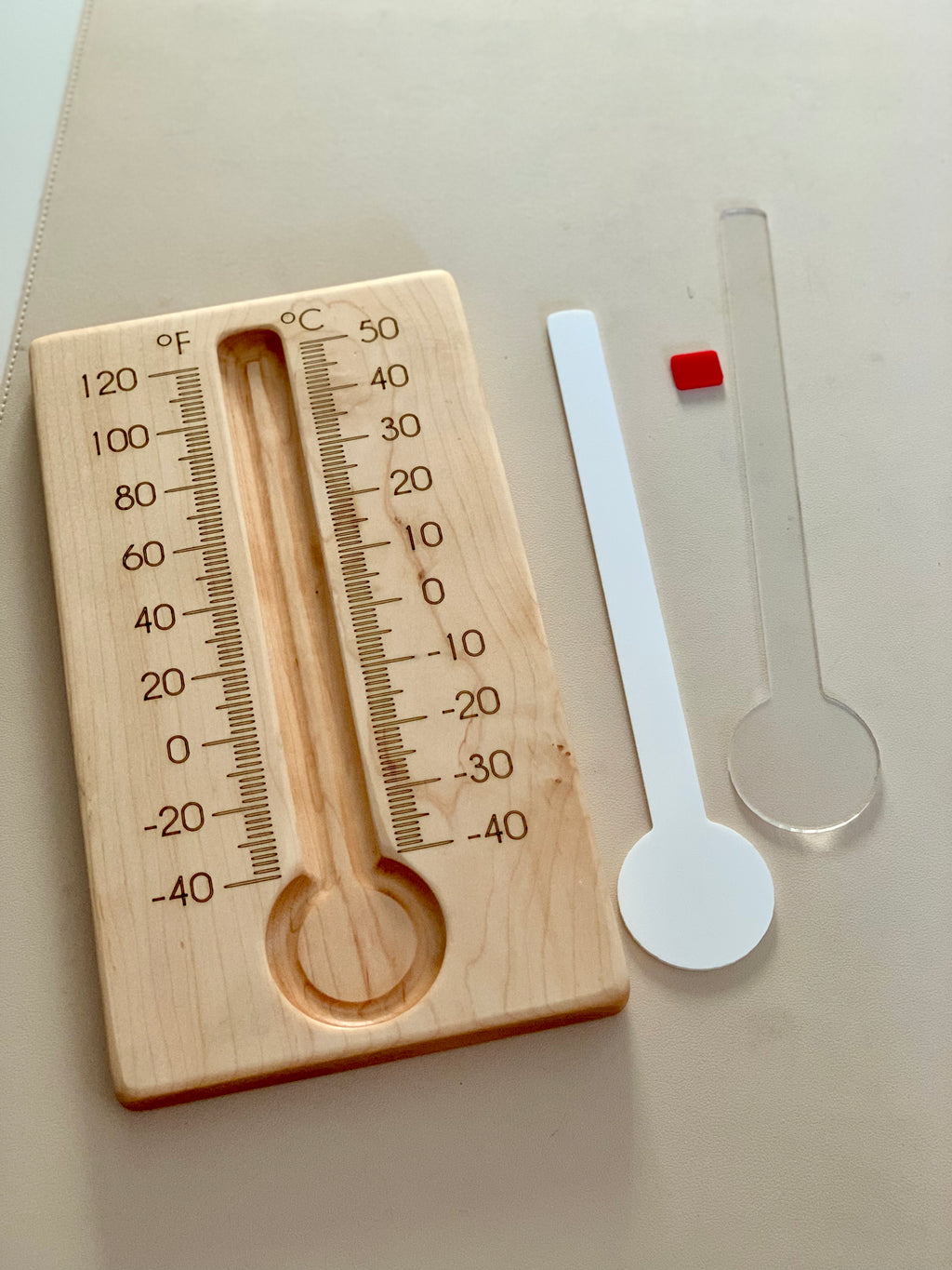 Wooden thermometer board with Fahrenheit and Celsius scales next to separate clear and white plastic thermometer inserts on light surface