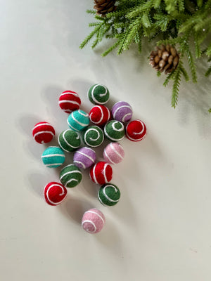 Assorted colorful felt balls with white swirl patterns scattered on a white surface near pine branches and cones