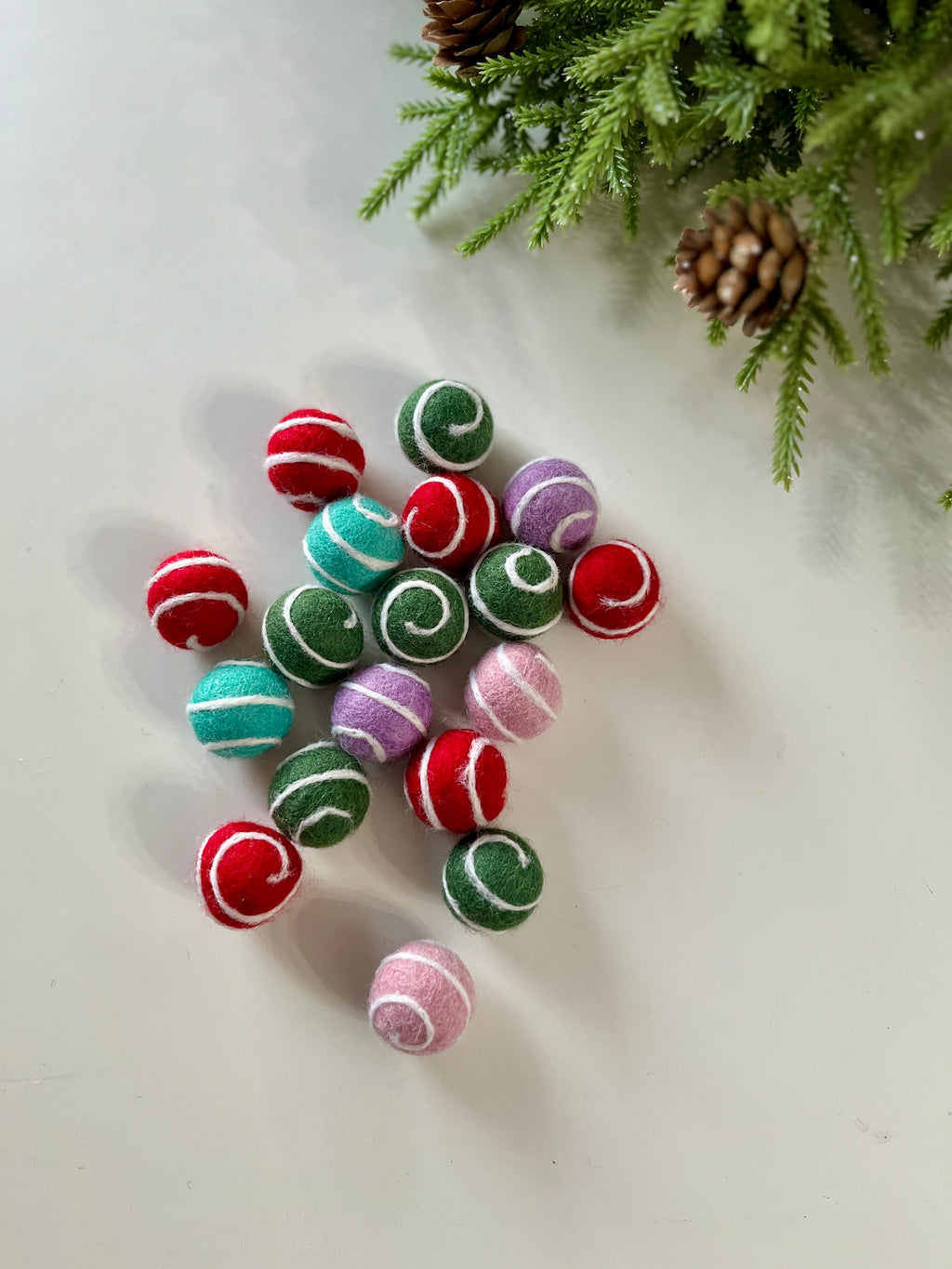 Assorted colorful felt balls with white swirl patterns scattered on a white surface near pine branches and cones