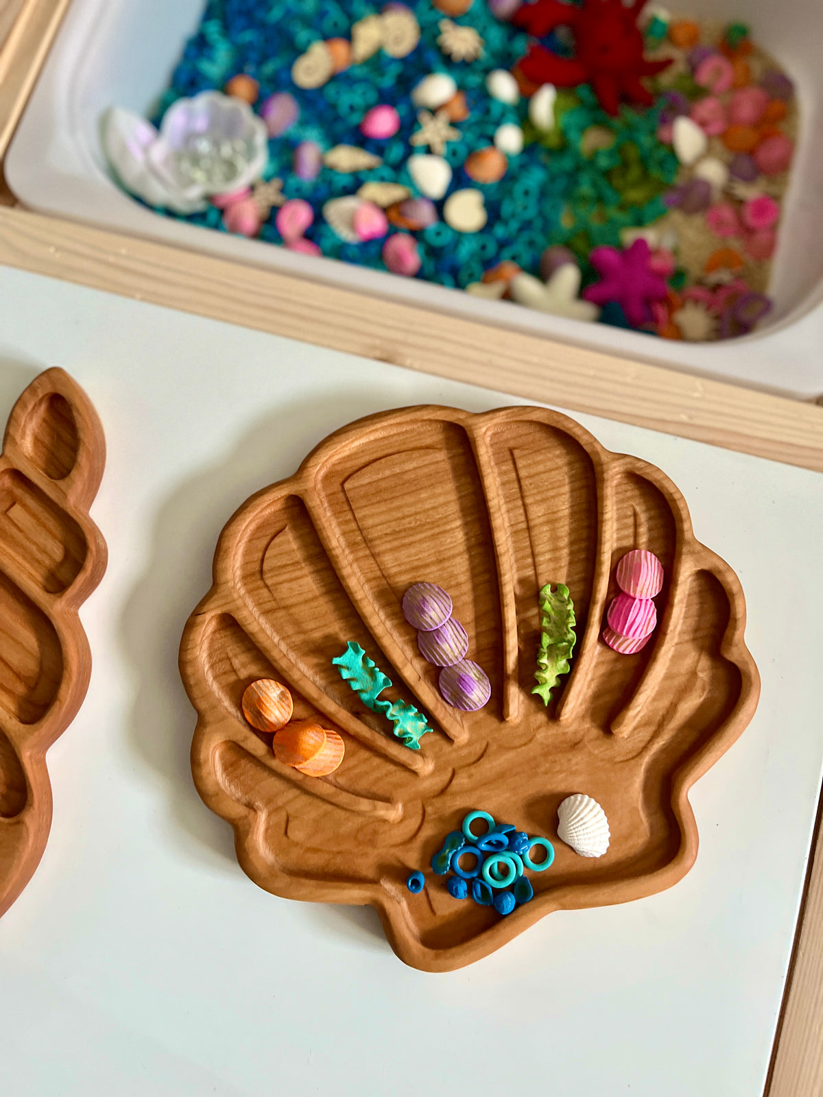 Wooden shell-shaped tray holding colorful felt seashells, green coral pieces, blue ring beads, and a white shell on a white surface