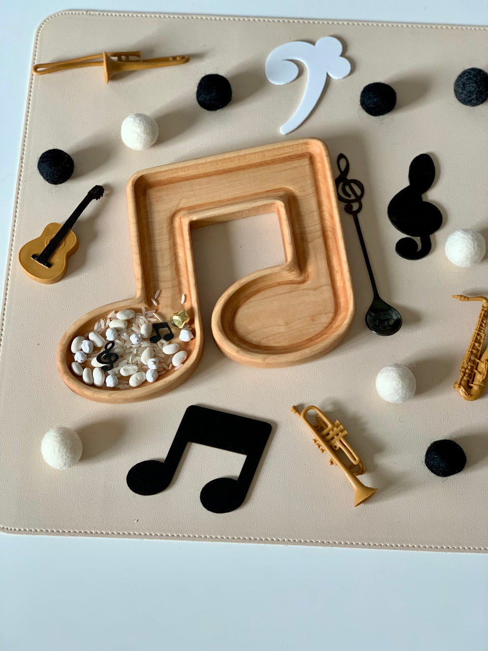 Wooden musical note tray with white beans and small black music tokens surrounded by miniature instruments and felt balls on light surface