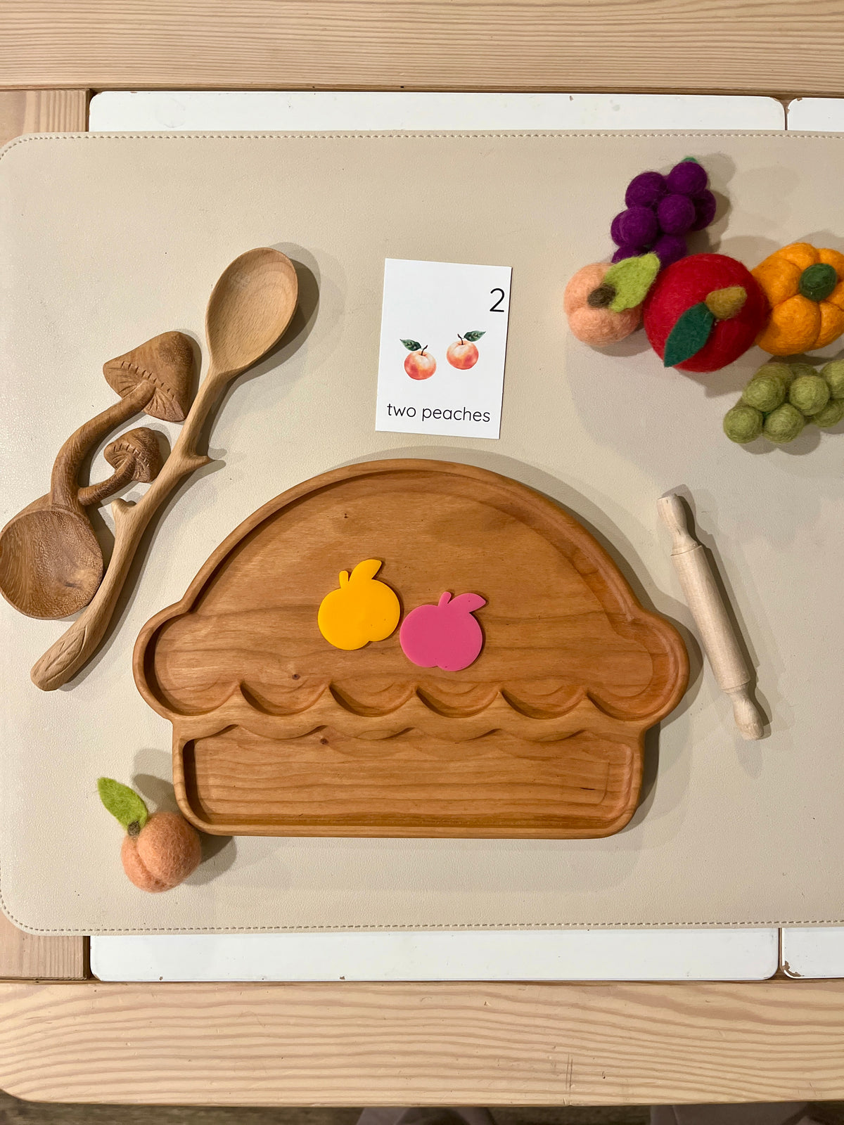 Wooden pie-shaped tray with two colorful peach-shaped cutouts, wooden utensils, and felt fruit on beige surface
