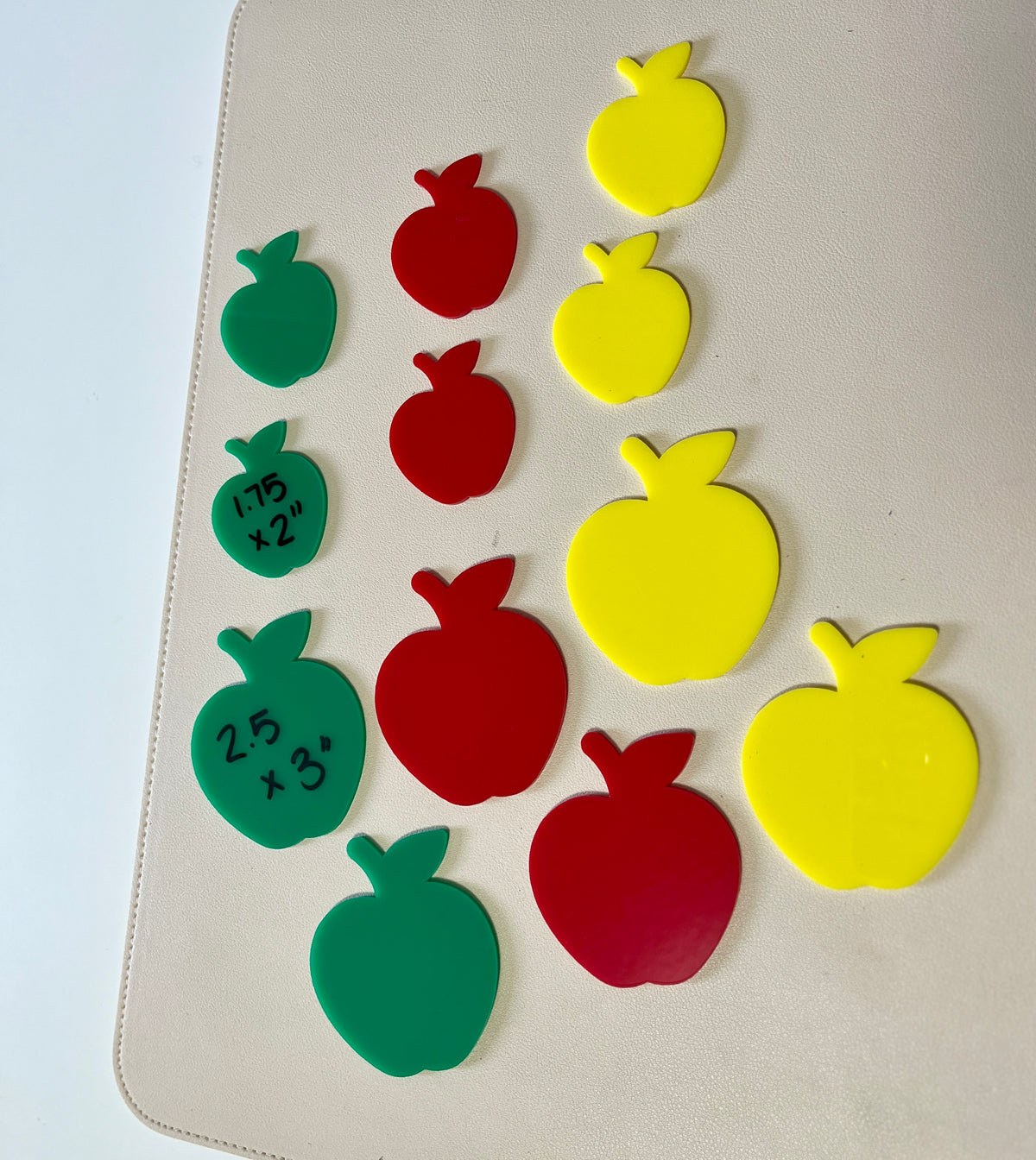 Green, red, and yellow apple-shaped plastic cutouts in various sizes arranged on a beige surface