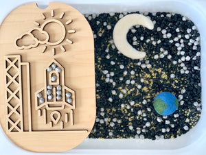 Wooden rocket puzzle with sun and clouds on a tray beside a sensory bin with black, white, and gold beads plus felt Earth and moon shapes