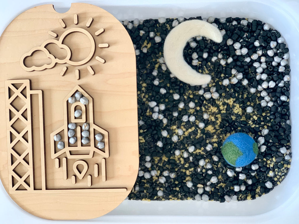 Wooden rocket puzzle with sun and clouds on a tray beside a sensory bin with black, white, and gold beads plus felt Earth and moon shapes