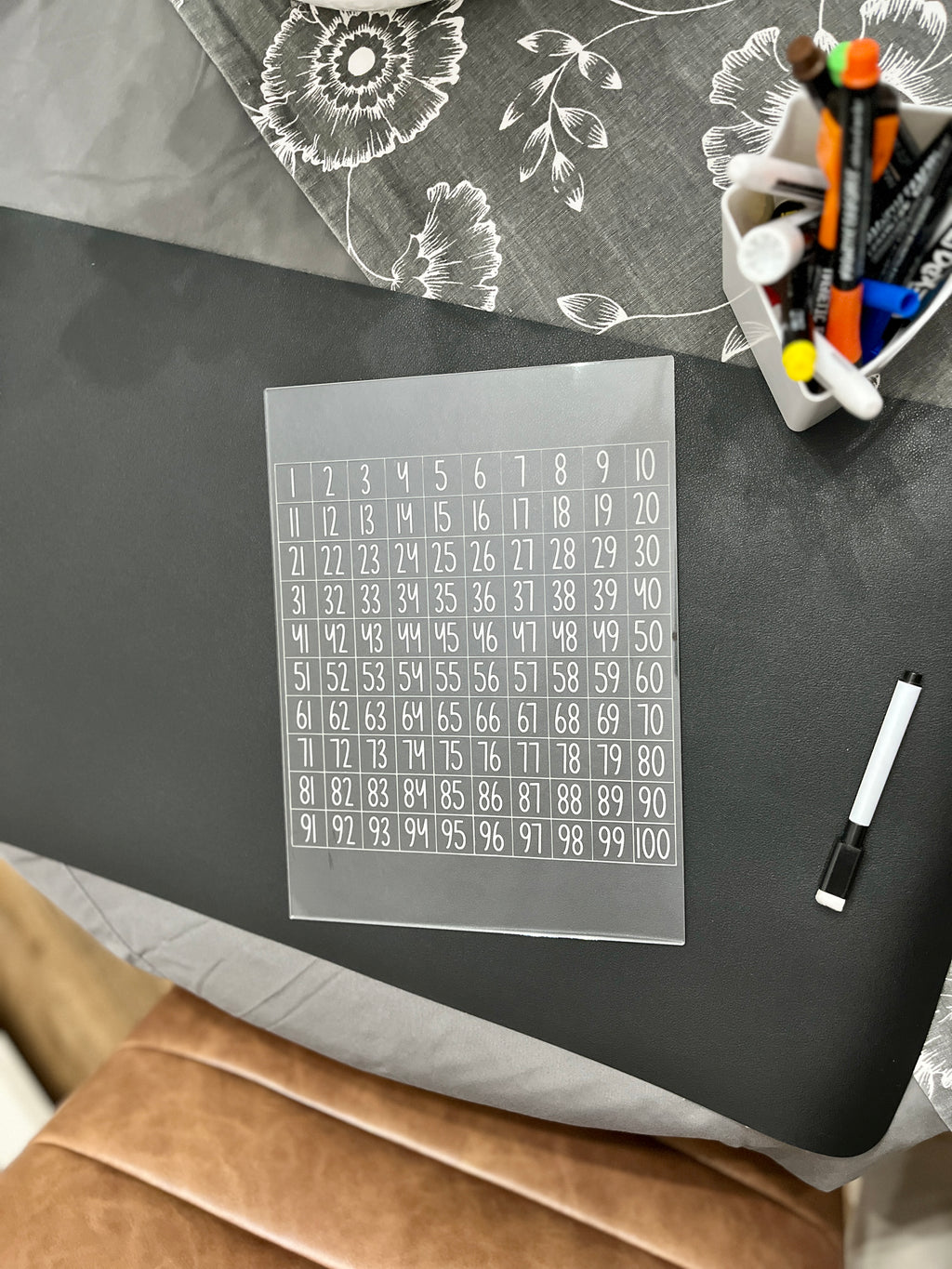 Transparent acrylic sheet with numbered grid 1 to 100 resting on black surface near whiteboard markers and patterned fabric
