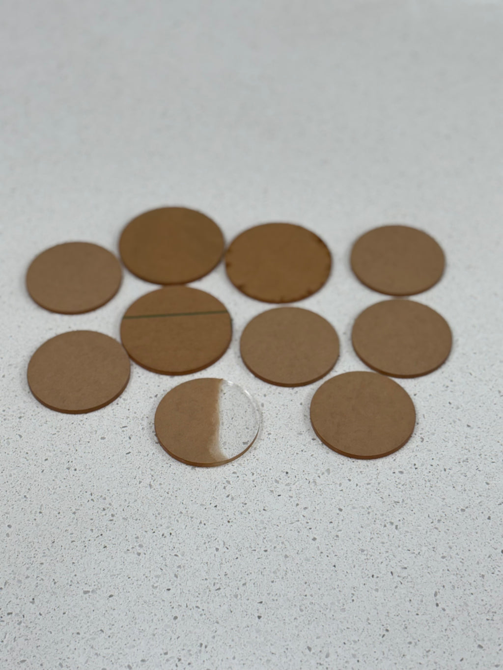 Ten round brown felt pads and one half-clear adhesive circle arranged on a light speckled surface