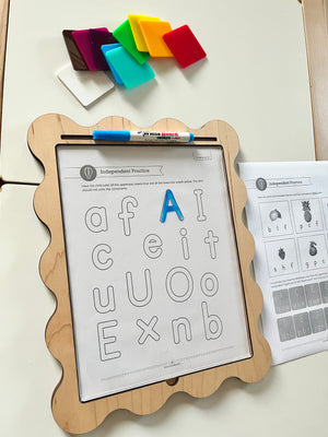 Wooden-framed activity board with scalloped edges holding a clear sheet over alphabet practice worksheet and colorful square tiles nearby
