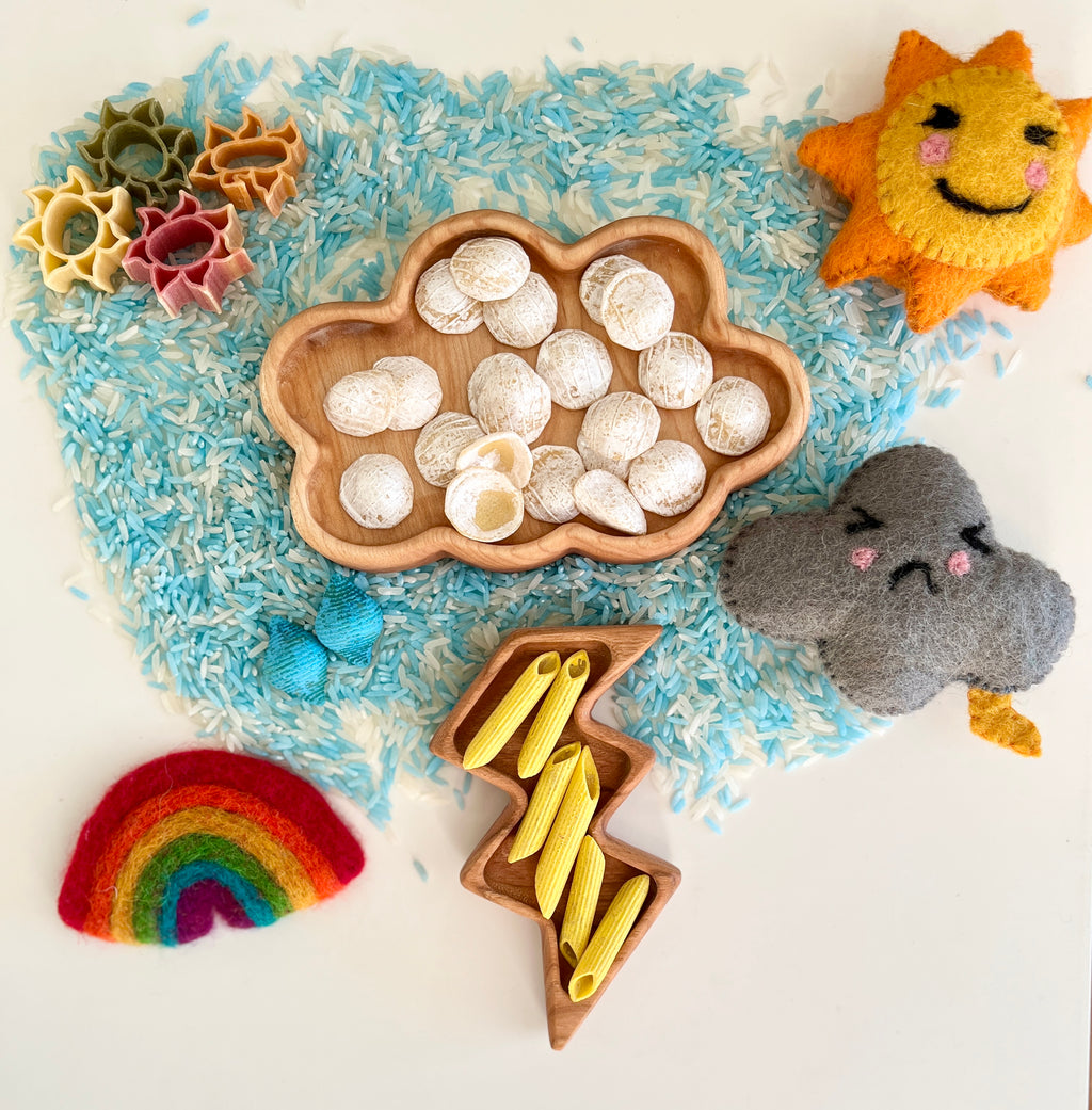 Wooden trays shaped like cloud and lightning filled with dried pasta and shells surrounded by felt sun, cloud, rainbow, and blue rice grains